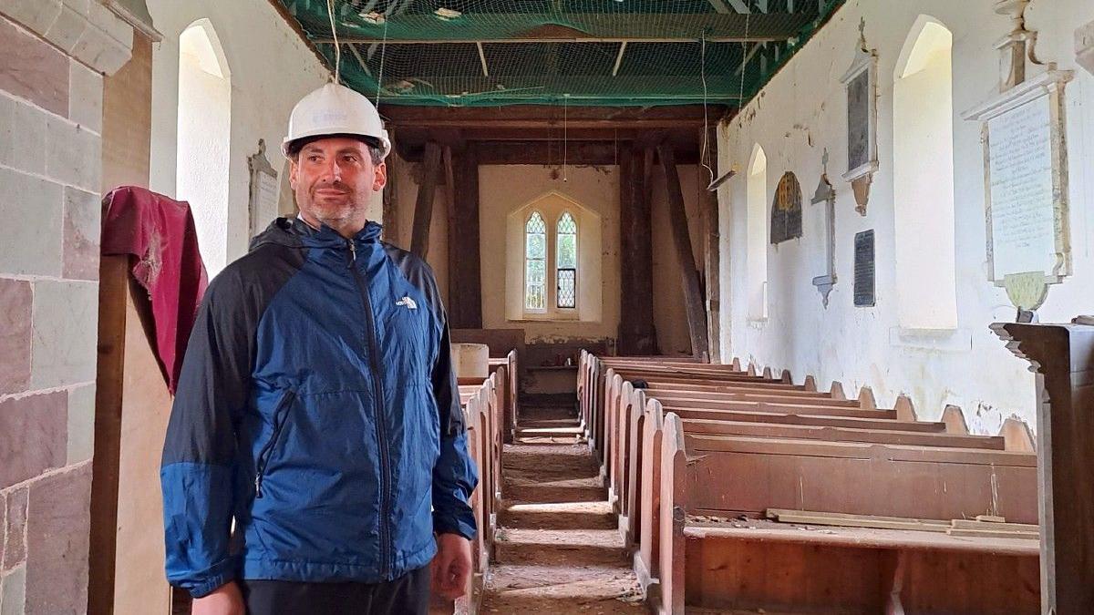 Sam Pratley stands near the pews in the church. There is debris on the benches and on the floor. The walls are marked. Church carvings are still on the wall.