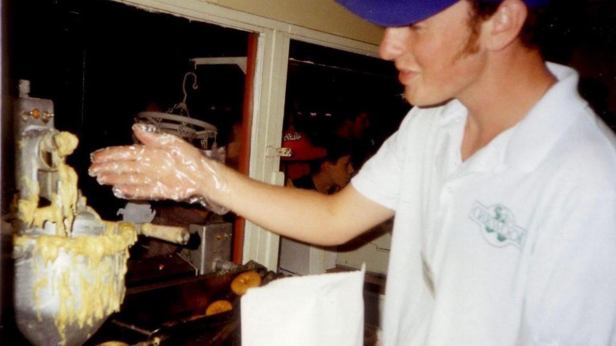 Richard working at food counter. He is wearing a blue cap and white polo shirt, with a green Oakwood logo on it. He is holding his arm out, and wearing a plastic glove. In the left hand side an electric mixing bowl can be seen with donut batter spilling over the side. Ring donuts can be seen in the background in another piece of equipment. 