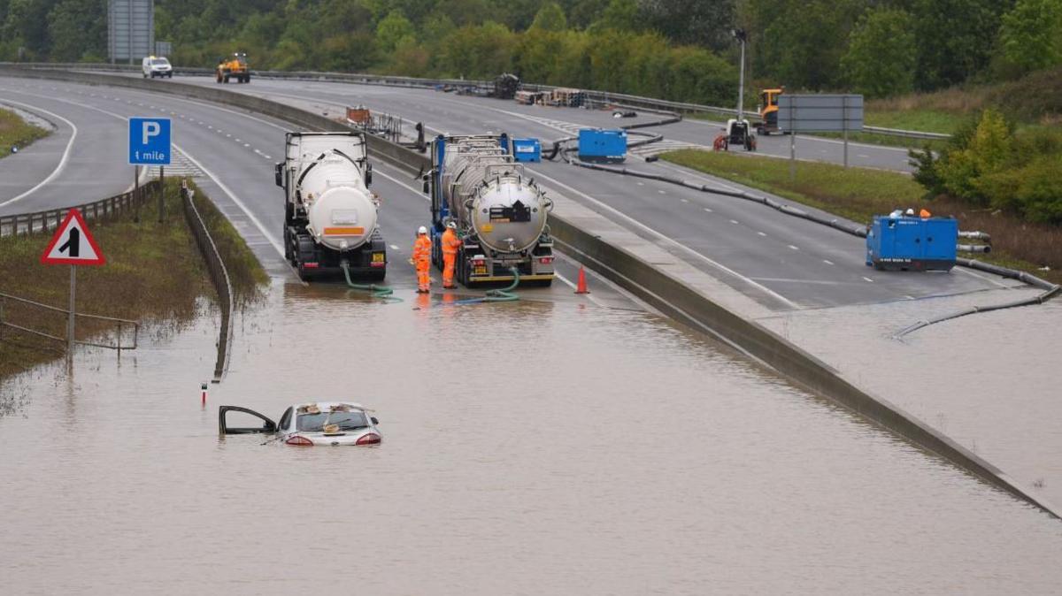 A car submerged in the floodwater covering the dual carriageway, with pipes trailing from two tankers and two workmen standing by