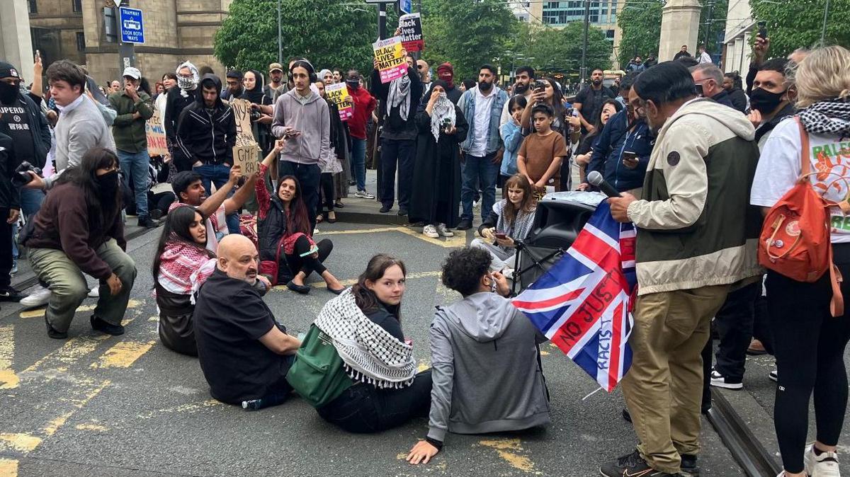 Protesters sit on the ground in Manchester city centre, surrounded by other demonstrators holding placards. 