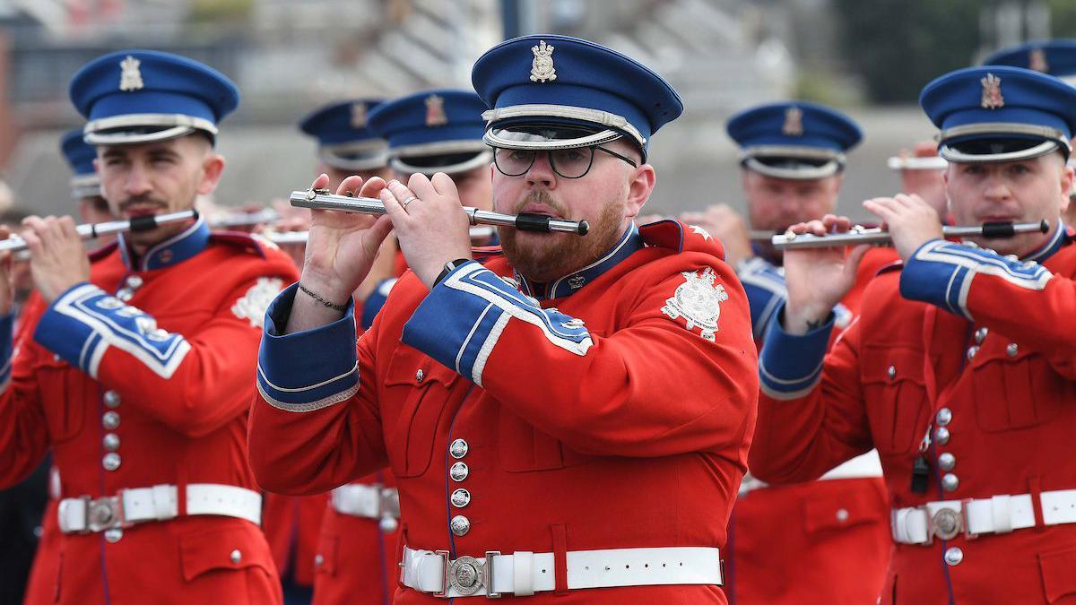 A number of men wearing red jackets and blue hats playing flutes. 