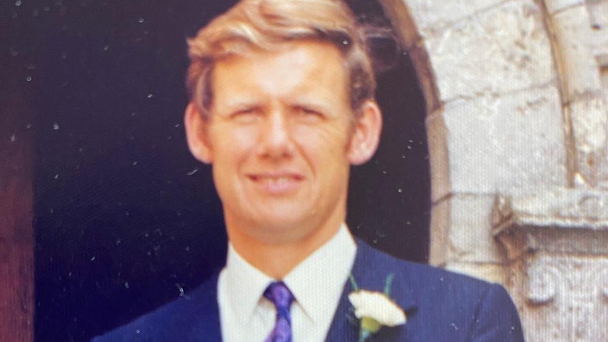 Gill Punt's father, Mike, stands outside a church and is wearing a blue suit, with a buttonhole attached to the lapel of his jacket. He has short blonde hair and is smiling.
