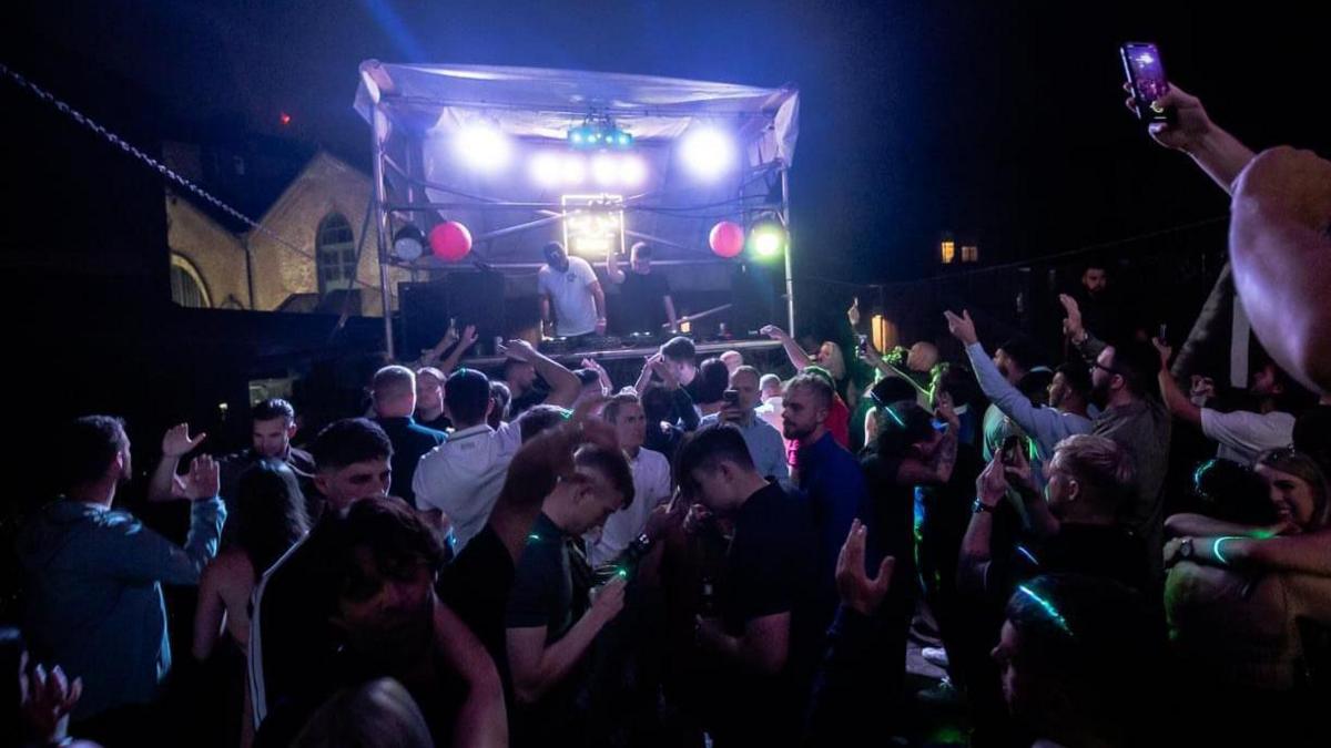 Dozens of people stood outside at a nighttime party, with a DJ on a heightened stage at Vines Bar in Derby