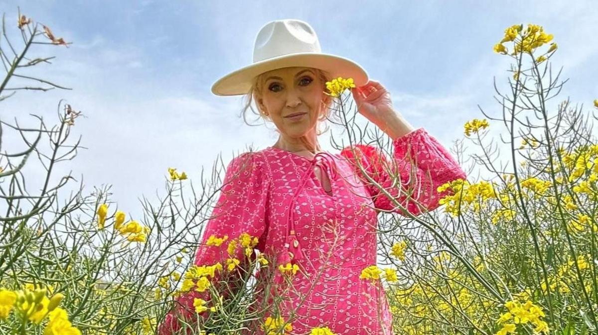 A woman wearing a pink dress and a hat. She is pictured in a field with yellow flowers around her.