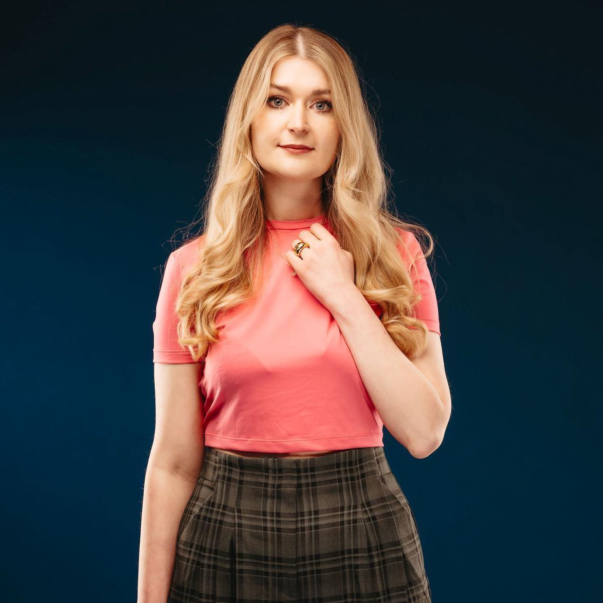 Josie Duncan is wearing a pink top and a tartan skirt. She is looking into the camera and is pictured against a dark blue background.