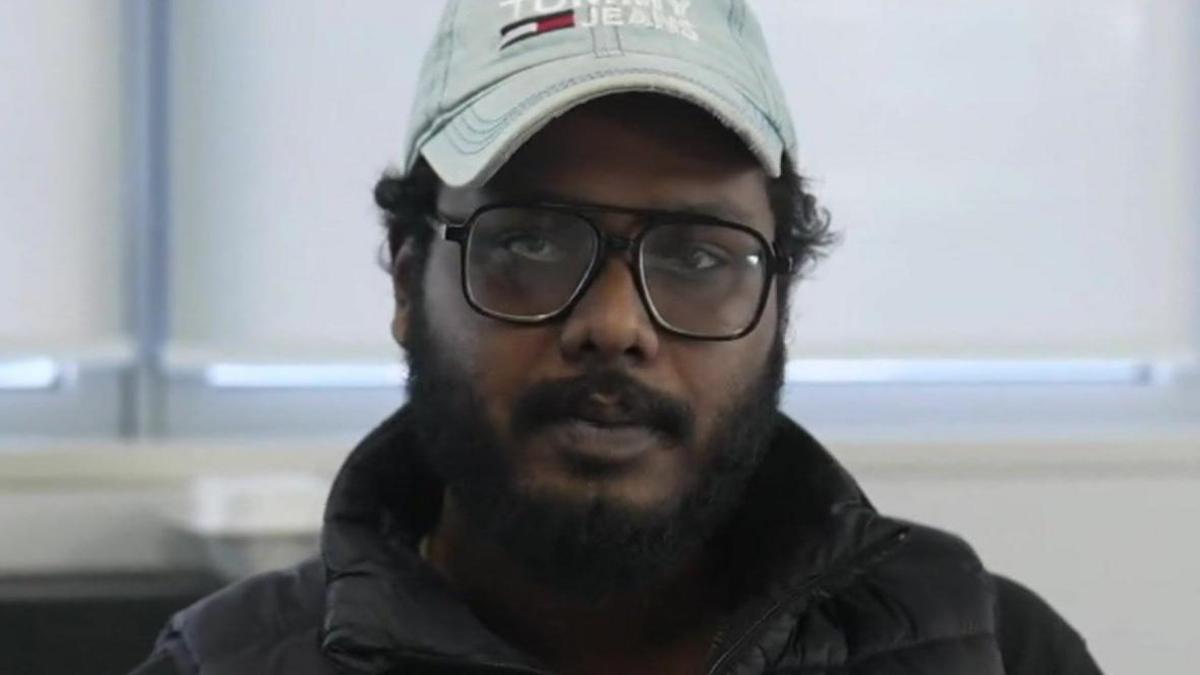 Nazmush Shahadat, a young man with dark hair, glasses and a beard in a black gilet and light blue cap. He is sitting in an office