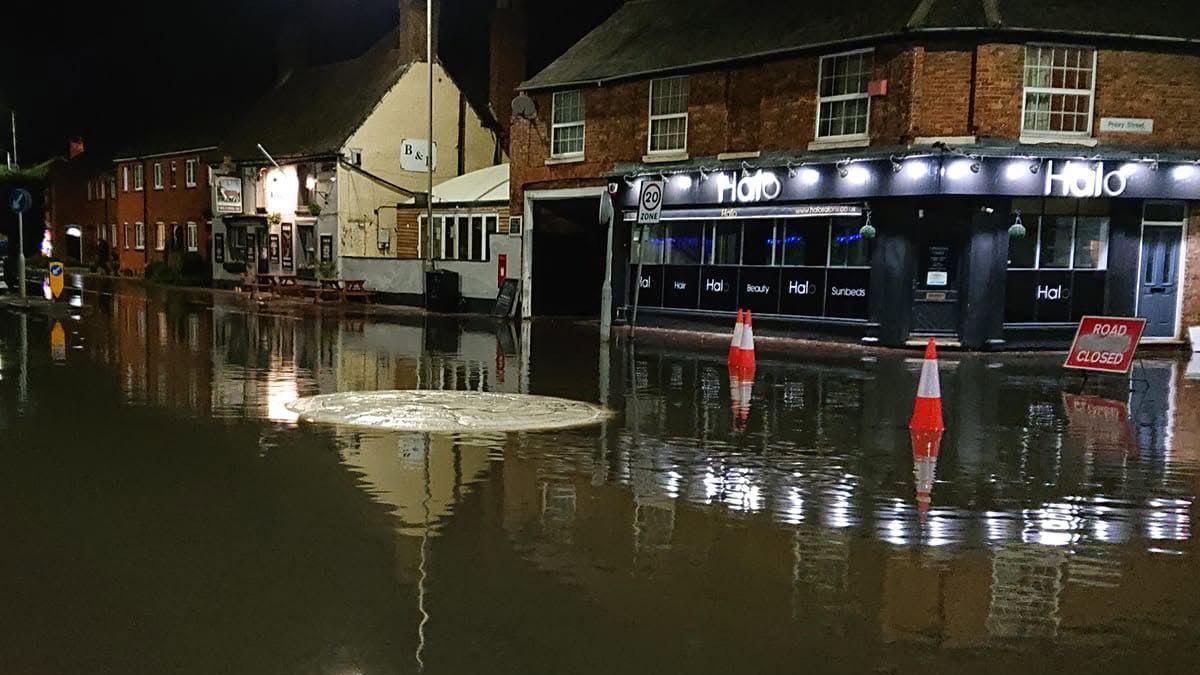 A flooded road in Newport Pagnell on Christmas Eve in 2020. A beauty salon called Halo can be seen on the corner of the junction of Tickford Street and Priory Street. It is surrounded by water; the white, painted top of a mini roundabout can just be seen poking out of the flood water. There are three traffic cones in the water and a 'road closed' sign at the top of Priory Street. It is night time and lights on the outside of Halo are on and are reflected in the water's surface. 