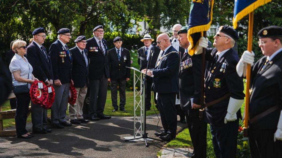 Men wearing smart jackets with war medals attached and berets stand in a row. Some hold wreaths made of paper poppies. A woman in a blue shirt and dark trousers stands next to them. They face the Mayor, who is reading a speech at a lectern. He wears a large gold chain draped across his shoulders. Two people holding flags upright can also be seen in the foreground.