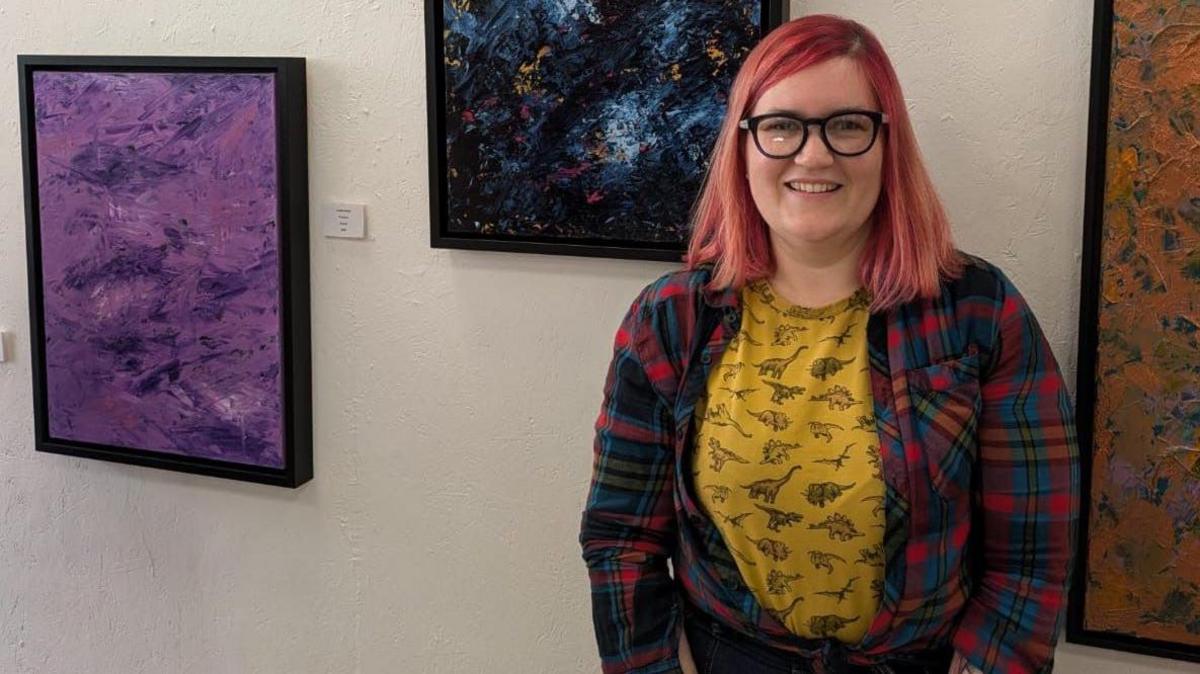 Louise Verity with long red hair in a yellow top and tartan shirt stands by a wall 