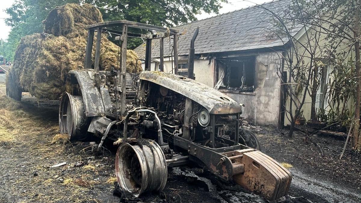 The chassis of a tractor after it being burned during the incident. There are charred hay bales just behind the tractor with a house seen on the right of the image. The house also displays clear damage caused by the fire as seen by the windows which have no glass or frames.