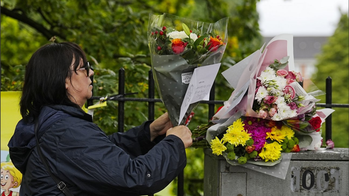 A woman in a navy blue waterproof coat, dark hair and dark glasses lays a bouquet of flowers on top of a cable box, where three bouquets have already been laid