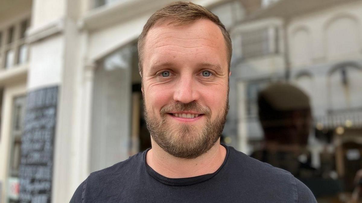 Ben Thomas  smiles at the camera, he has short brown hair and a beard covering most of his chin and wears a black T-shirt, standing in front of the bakery