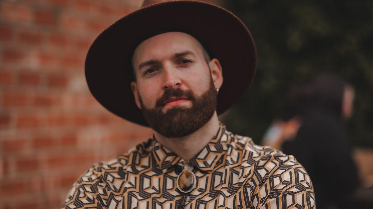 Tony Beacock. He has a well-trimmed beared and moustache. He is wearing a large-rimmed brown hat, and a patterened, buttoned-up light brown shirt with a matching lace tie.
