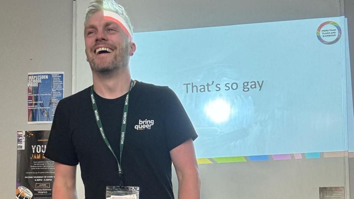 Ian Timbrell, wearing a black T-shirt with the logo 'bring queer', laughs as he stands in front of a whiteboard with the words 'that's so gay' projected on to it.