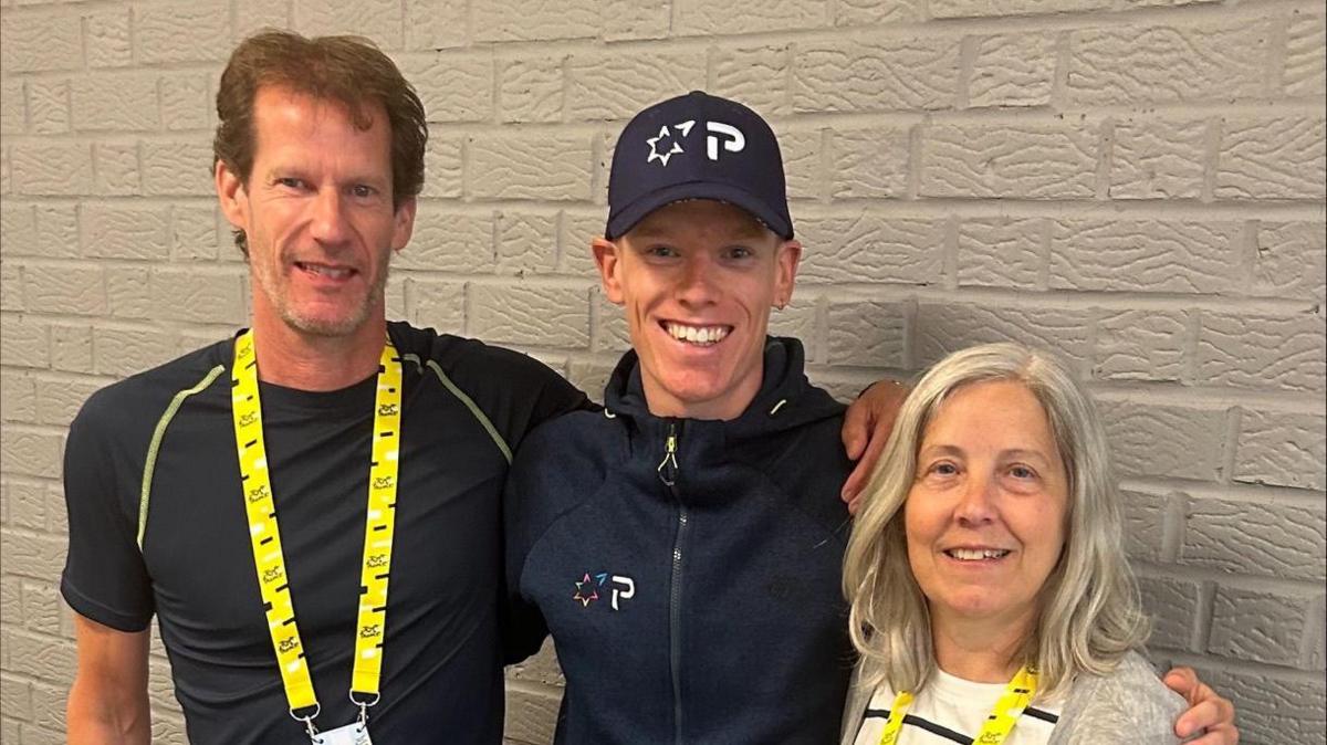 Two men in dark blue tshirts and a woman in a white and black striped shirt and grey cardigan are standing in a line with their arms around each other and are smiling at the camera. The man and woman on either end of the row are both wearing yellow lanyards while the man in the middle is wearing a cap