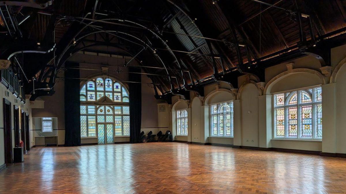 The Great Hall inside the building. The hall is empty and has stained glass windows and polished wooden floor.
