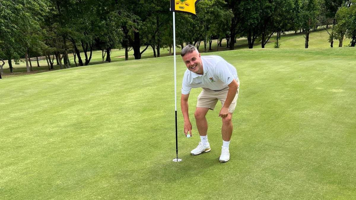 Vigurs wearing a white polo top and beige shorts bends over and picks out a golf ball from the hole on the green.