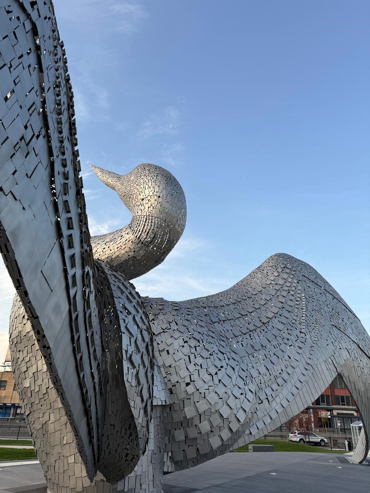 Rear side view of 'The Calling', large steel sculpture of a loon bird with its head thrown back and wings fully outstretched, surrounded by a paved viewing area.