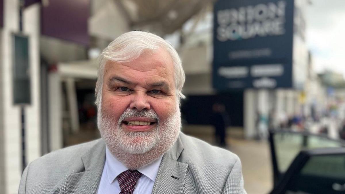 The Conservative leader of Torbay Council, David Thomas, wearing a white shirt and a burgundy and white spotted tie and a grey blazer with a blurred background showing a sign for Union Square shopping centre in Torquay.
