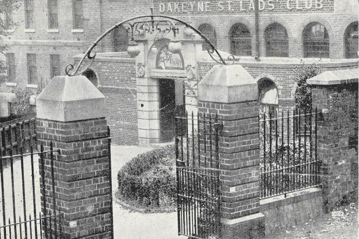 Vintage photograph showing two brick pillars forming the entrance to a building which has a sign saying Dakeyne Street Lads Club