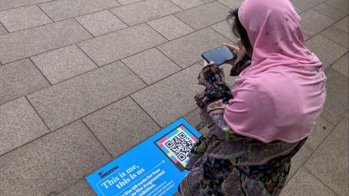 A woman in a floral dress and a pink hijab is looking at her phone which is pointed at a blue sign on the floor which bears a QR code and the writing, This is me, this is us.