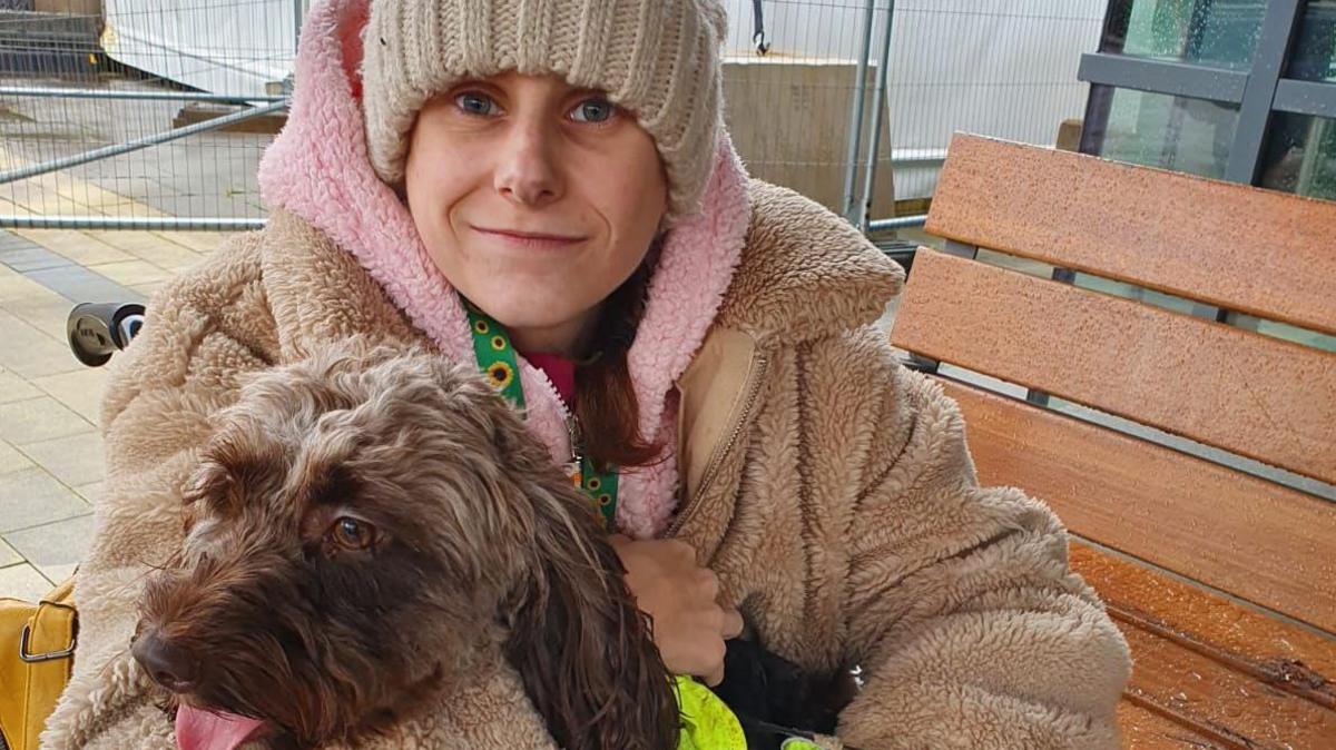 Molly Leonard pictured with a brown long-haired dog. Molly is wearing a cream knitted hat with a pink, fluffy hooded jumper and a beige fluffy jacket on top, and is sitting on a wooden bench. 