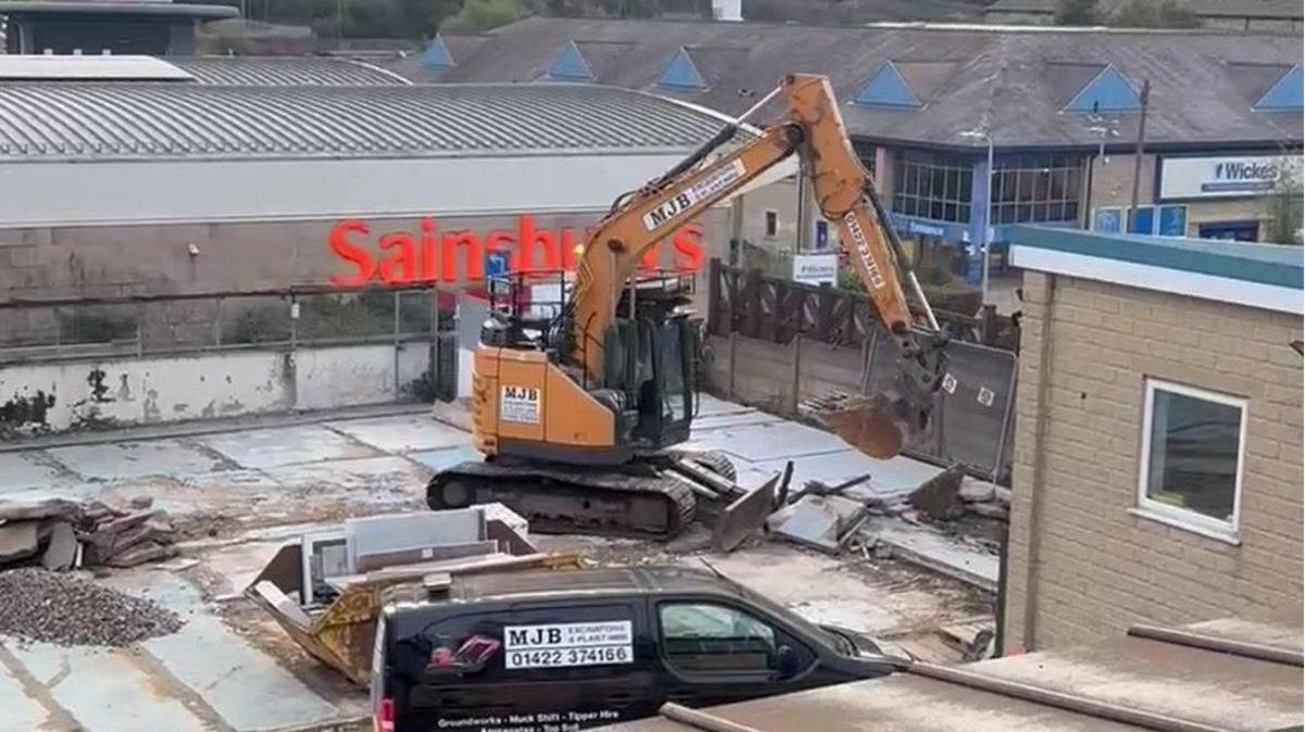 A building site with two vehciles parked alongside a skip and the remains of a demolished building
