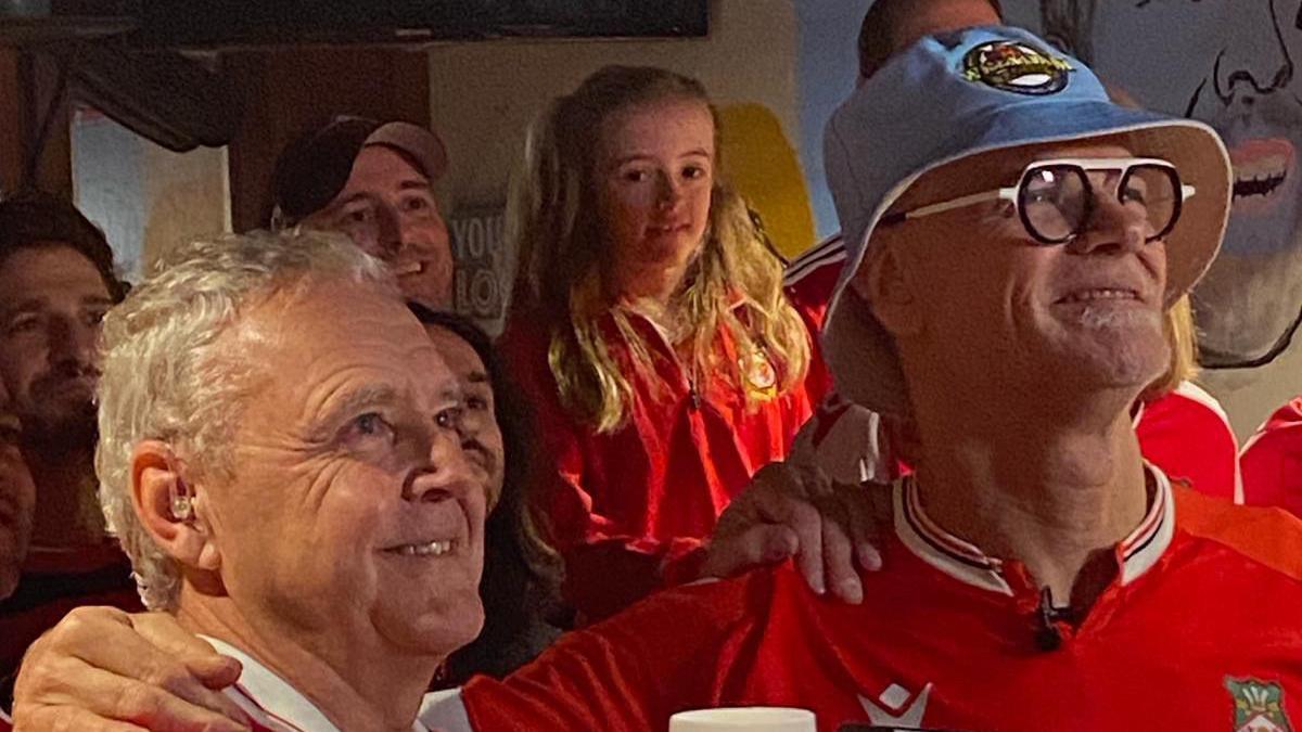 Mike and Dieter surrounded by Wrexham fans looking up most likely at a television as a Wrexham game is on. They are wearing Wrexham tops. And Dieter has a blue hat on.
