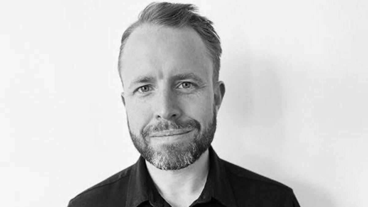 A head and shoulders black-and-white photo of a man wearing a black shirt, with short hair and a beard, smiling at the camera.