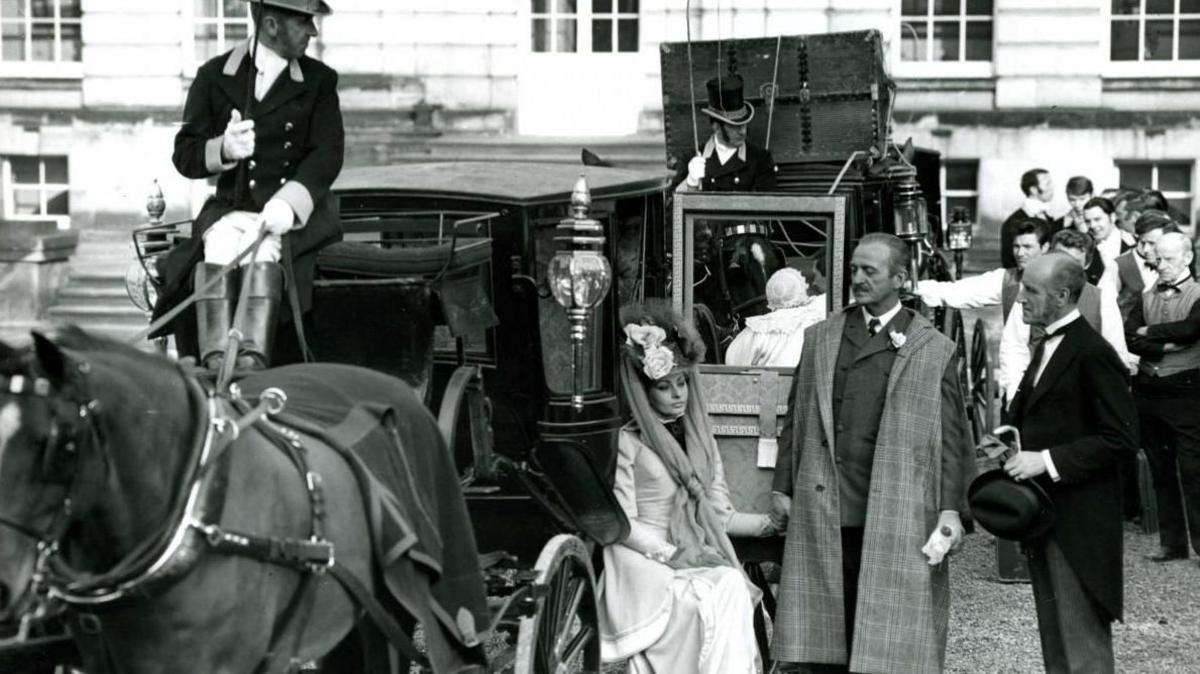 David Niven and Sophia Loren on set in Lady L. Loren is sat on a carriage being towed by horses.