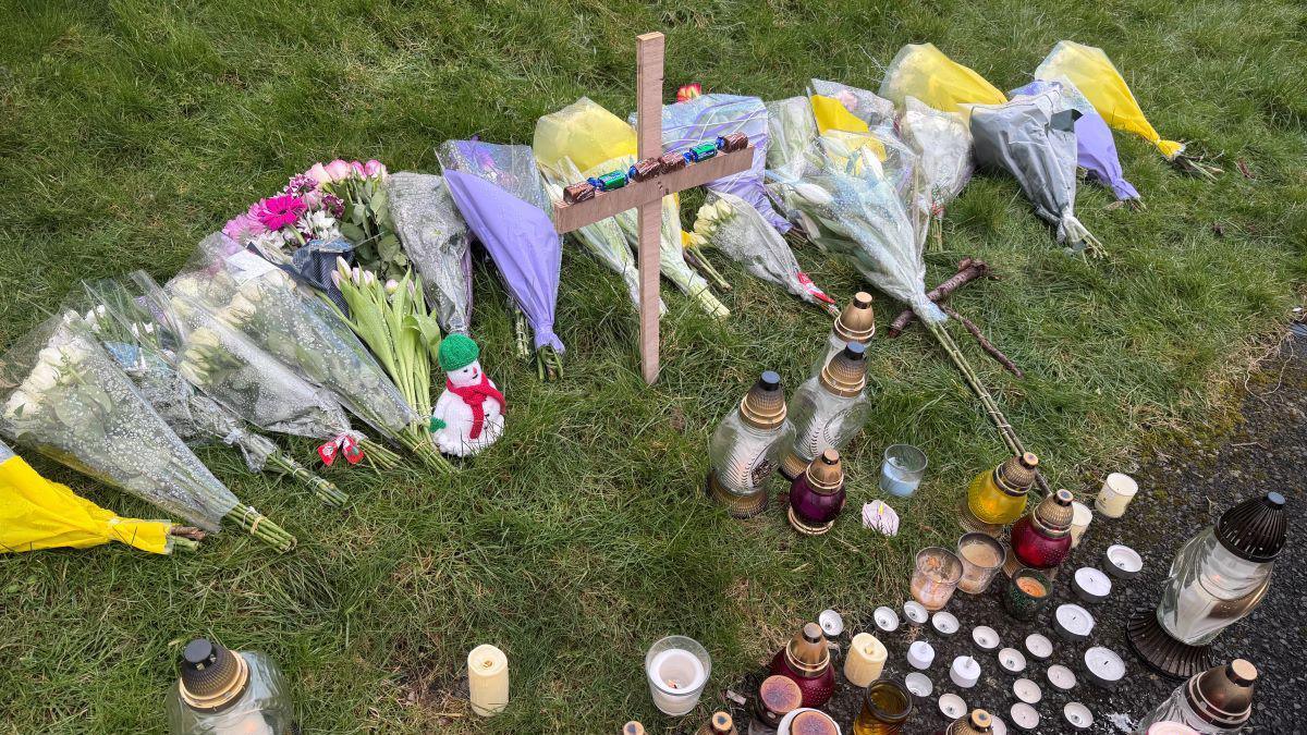 A wooden cross in green grass with 19 bouquets of flowers and various different coloured candles surrounding it.