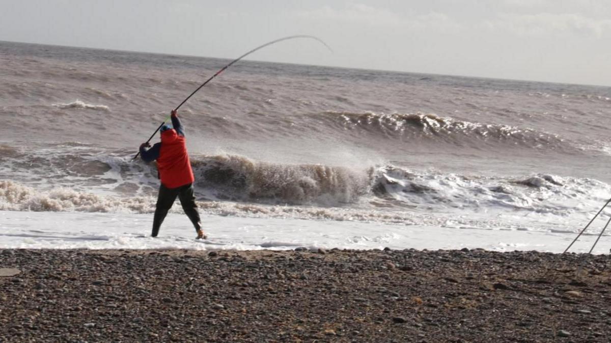 A man can be seen on a stony beach holding a fishing rod above the surf of a grey-brown sea near the shoreline. He has his back to the camera and is wearing a red jacket.