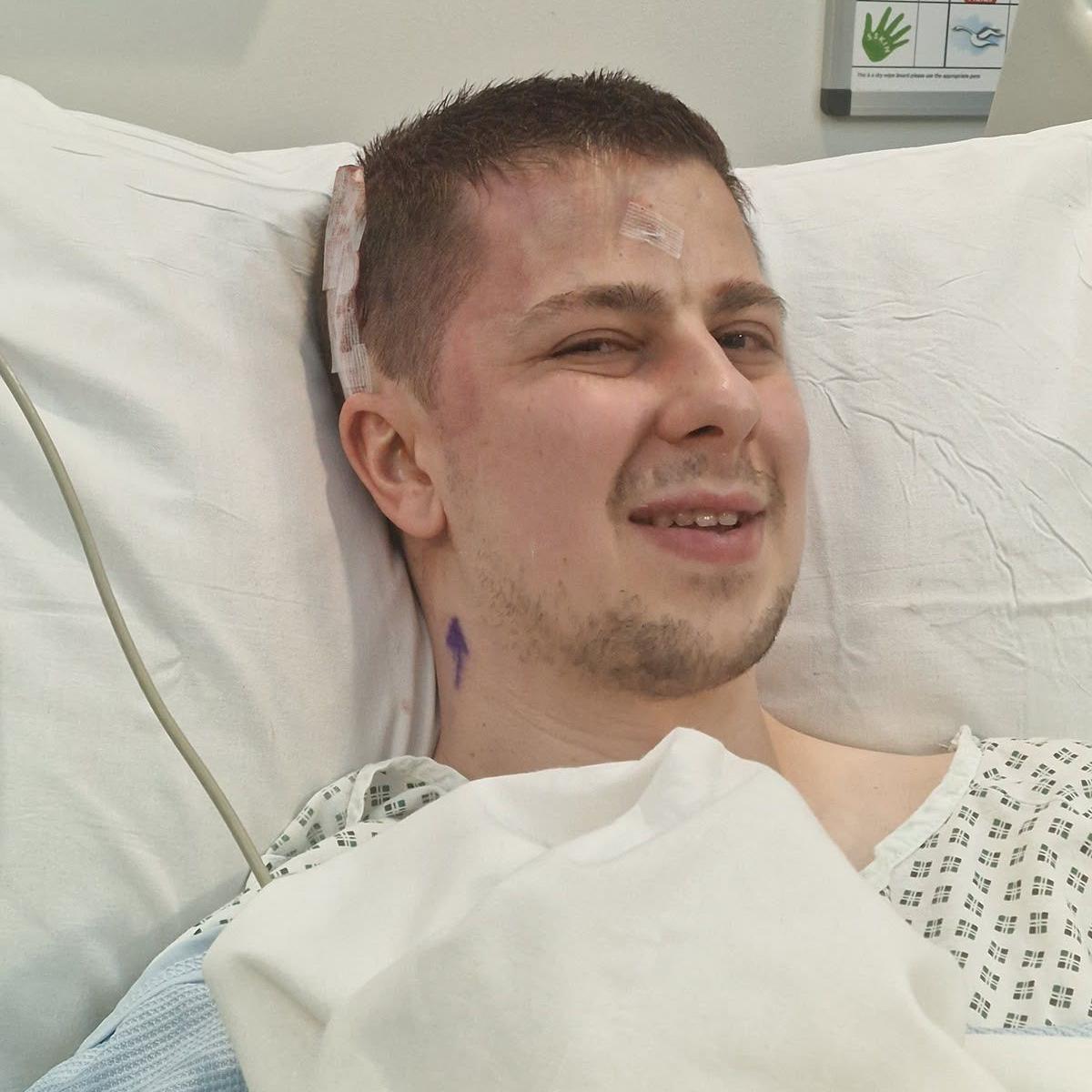 Joshn wearing a hospital gown and lying in a hospital bed with white bedding. He has a piece of surgical tape along one side of his head and is looking at the camera and half-smiling.