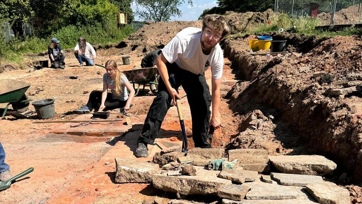 A man wearing a white t-shirt and black trousers digging at the site of the former Tudor mansion site