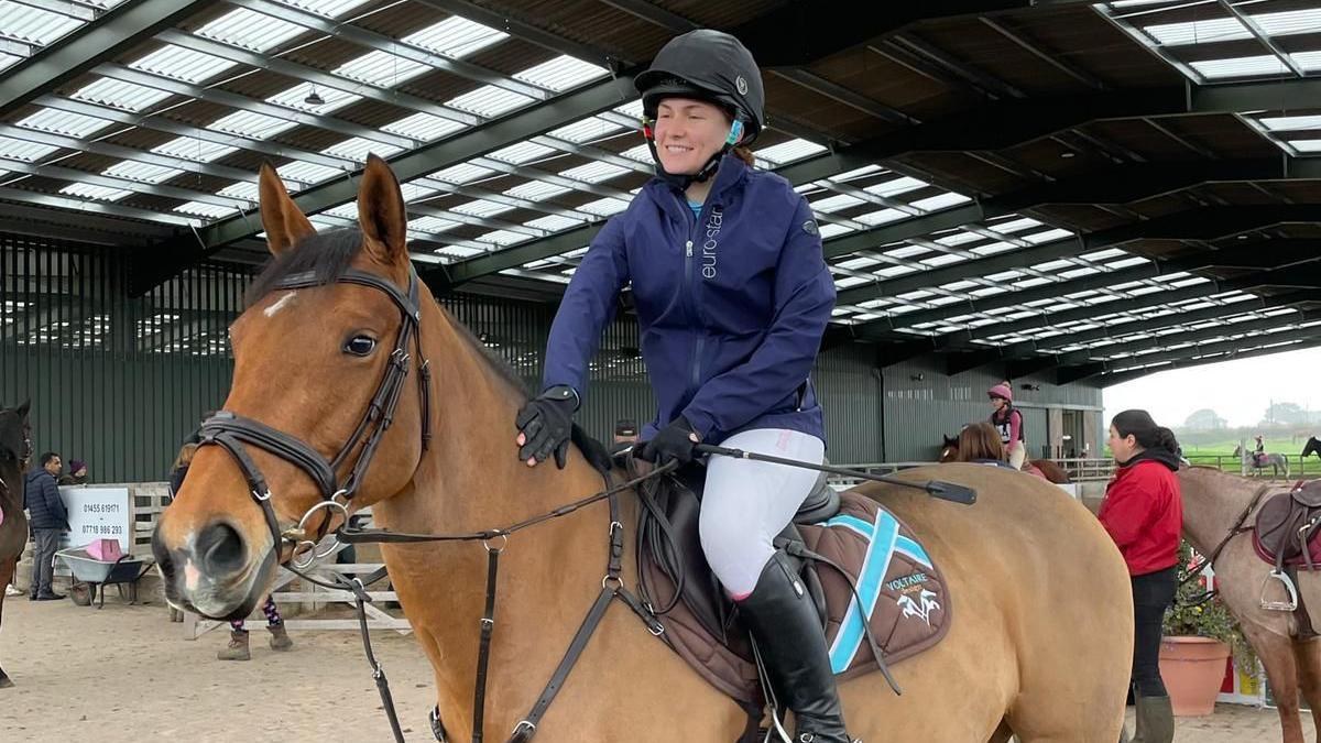 Millie Tingey sits on her bay horse in a riding school, surrounded by other people on horses and spectators, in English horse riding attire. She is smiling as she pats her horse, Baby, on the neck.