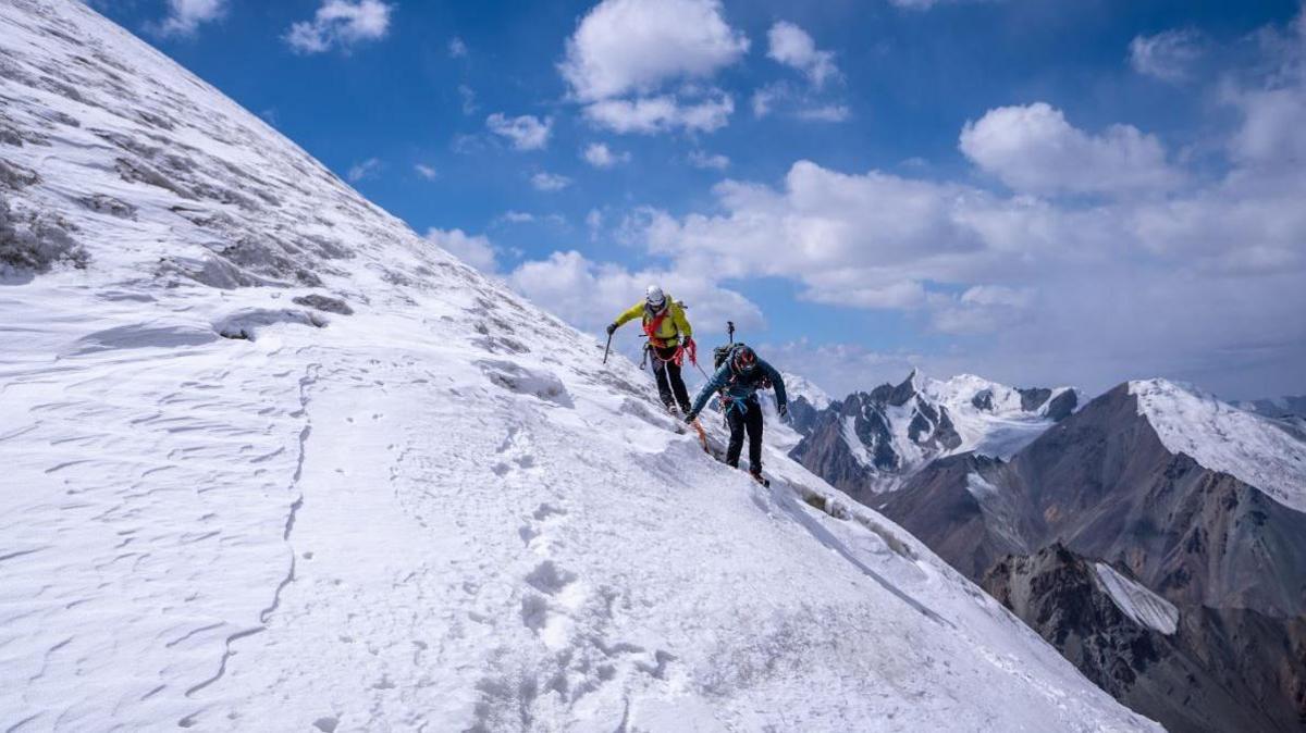 Two people wearing helmets and colourful jackets, using hiking poles to traverse down a steep snowy slope on the side of a mountain. They are extremely high up and in the background you can see several other peaks in the mountain range, which are smaller and all covered in snow. the sky is a bright blue with lots of fluffy white clouds.