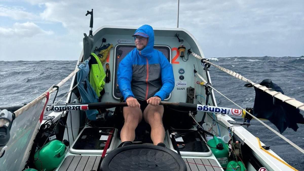 Dan Lewis in a blue coat sits on The Mayflower. He is rowing in the middle of the Atlantic. There are waves in the background around him. 