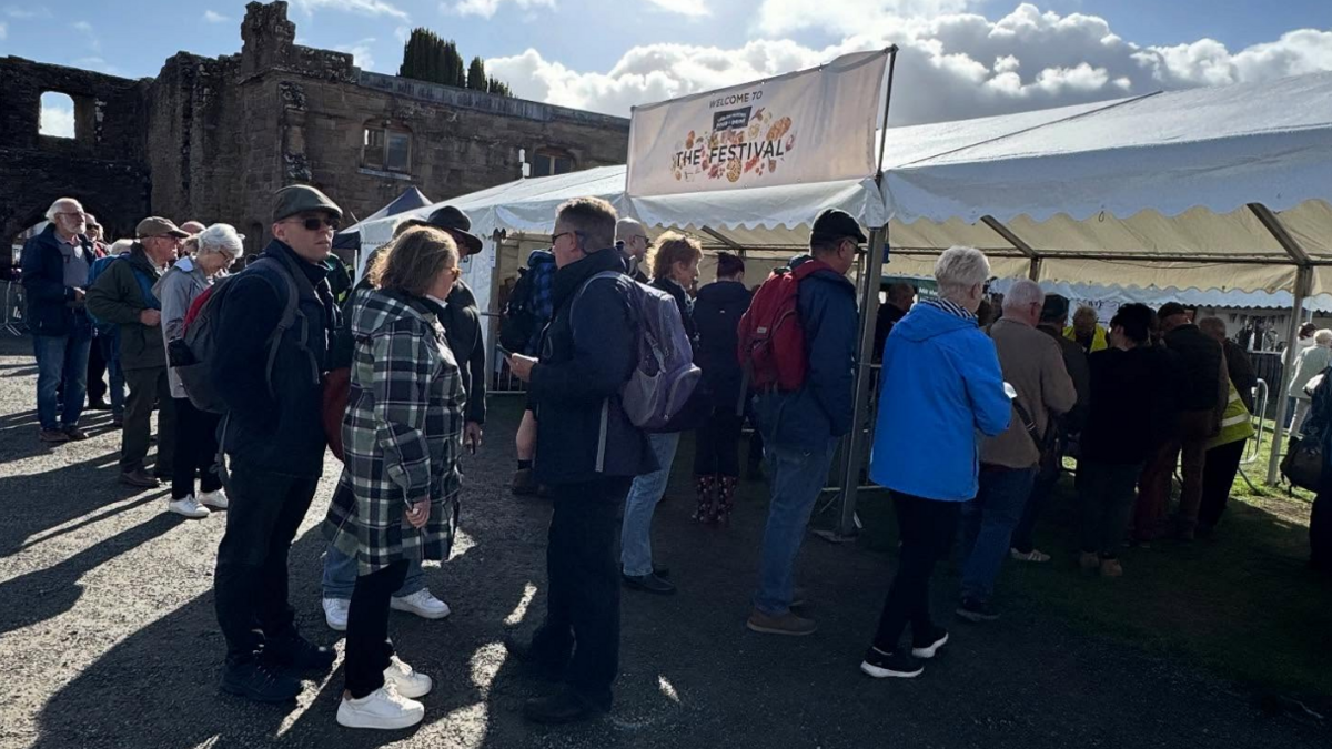 A crowd of people approaching ticket and bag checks under an open-sided white marquee with a "Welcome to the Festival" banner above. In the background, the castle's stone walls are visible.