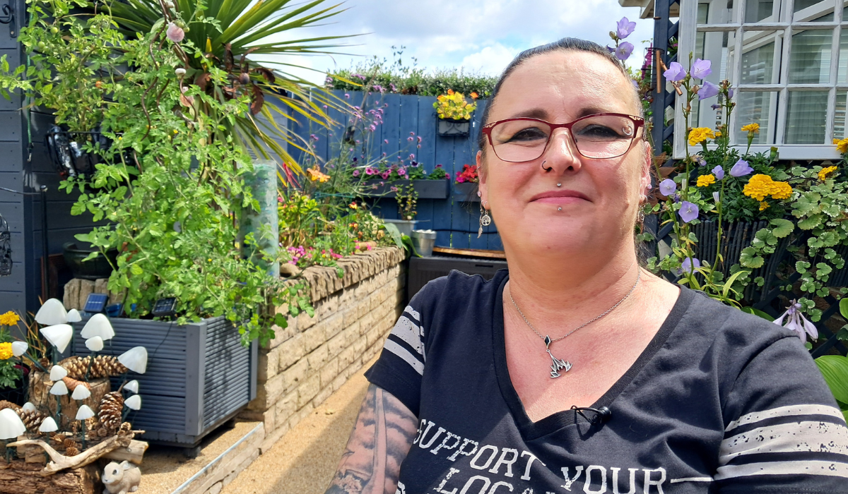 Victoria Coates is wearing a black t-shirt and sitting in a garden with flowers in the background.