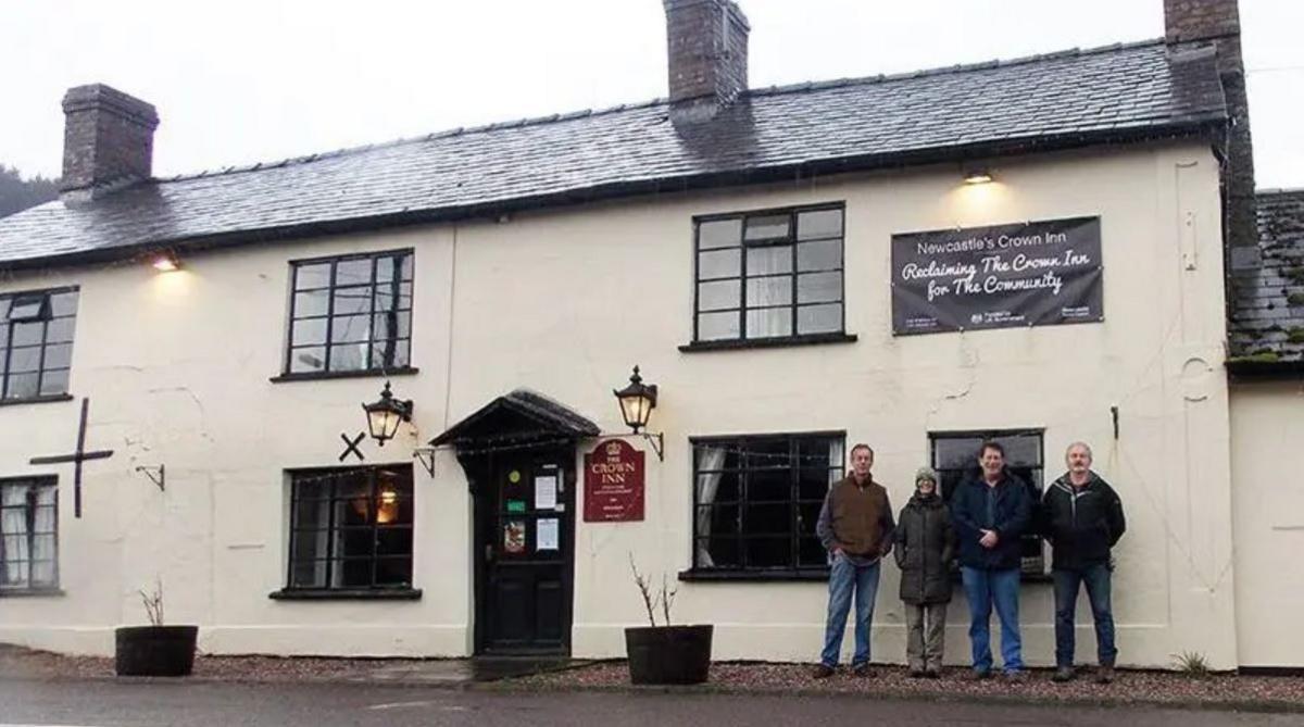 A white building with black framed windows and door, a black tiled roof and four people standing outside. There are two vintage lamps on the wall on each side of the entrance door. There is also a burgundy sign with Crown Inn written on it. Above the four people there is a black banner attached to the wall, saying "Newcastle's Crown Inn - Reclaiming the Crown Inn for the Community".