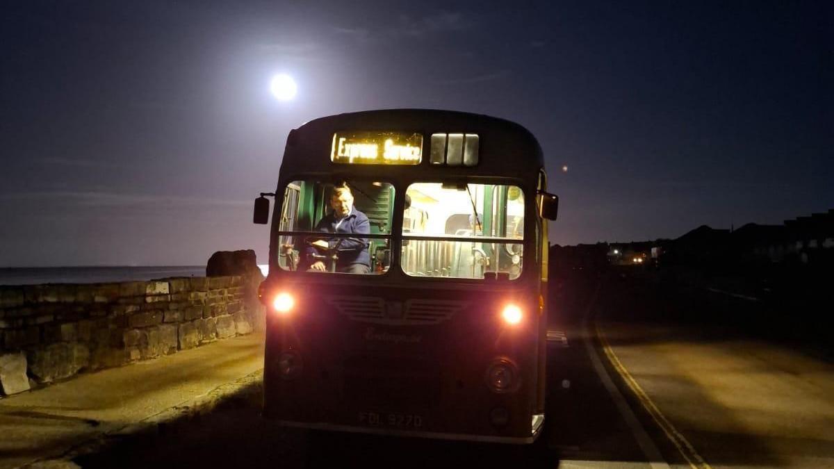 A bus in the middle of the road with the moon in the sky just to the rear of the bus