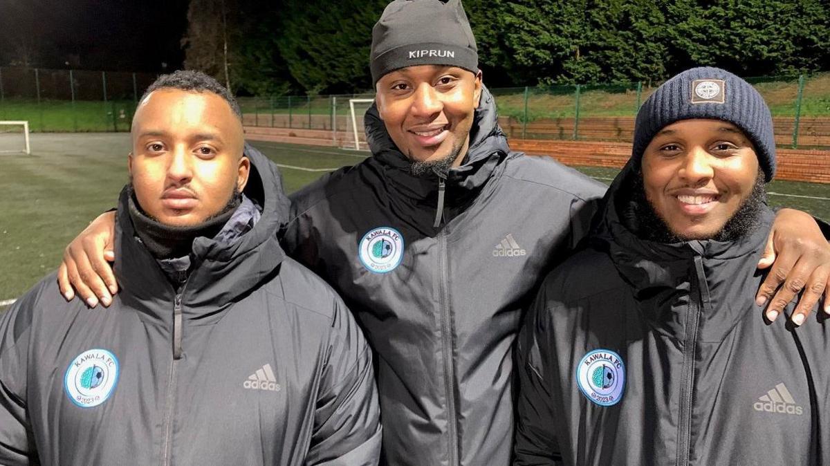 Three men face the camera, all wearing grey/black tops with a round badge with the words "Kawala FC" on with their logo. The one on the left has short brown, curly hair and does not smile. The one in the middle wears a black hat and smiles and has a short black beard. The one on the right smiles broadly, has a short black beard around his face and wears a blue woollen hat.