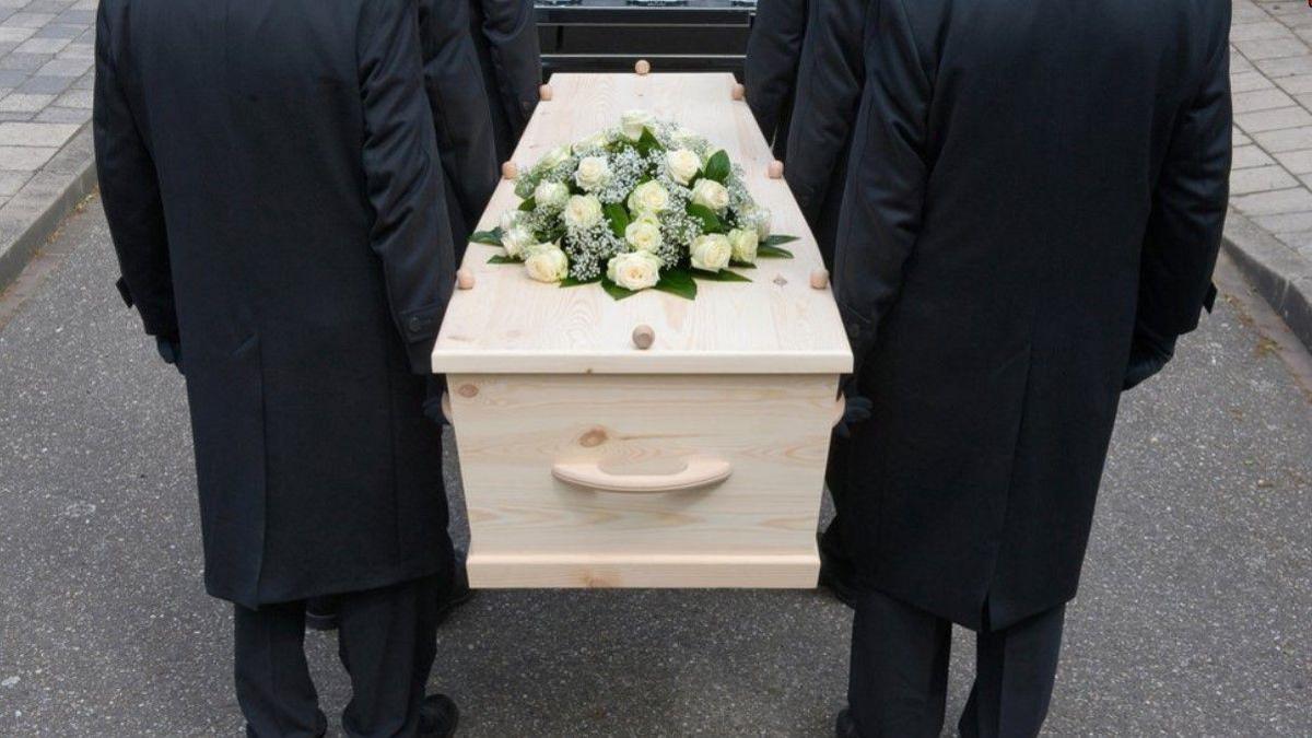 Three people on the left and three on the right, dressed in black and near a pavement, carry a coffin. Flowers are on top of the coffin. 