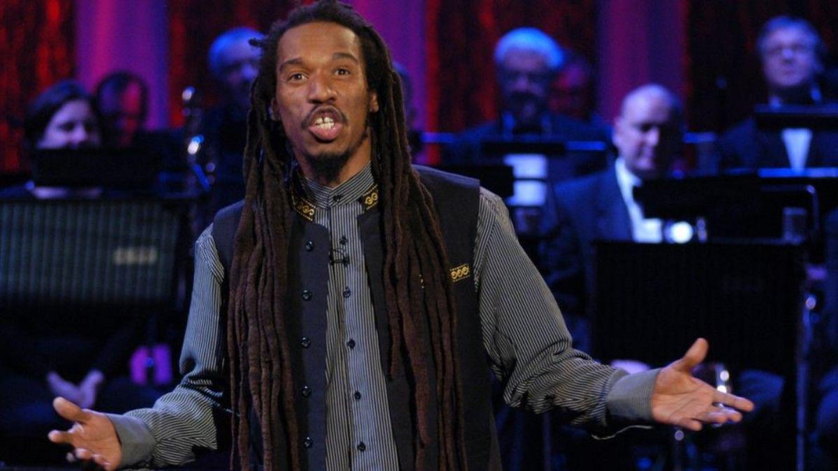 Benjamin Zephaniah, with dreadlocks, is holding both of his arms out, as he recites a poem. Musicians are in the background. 