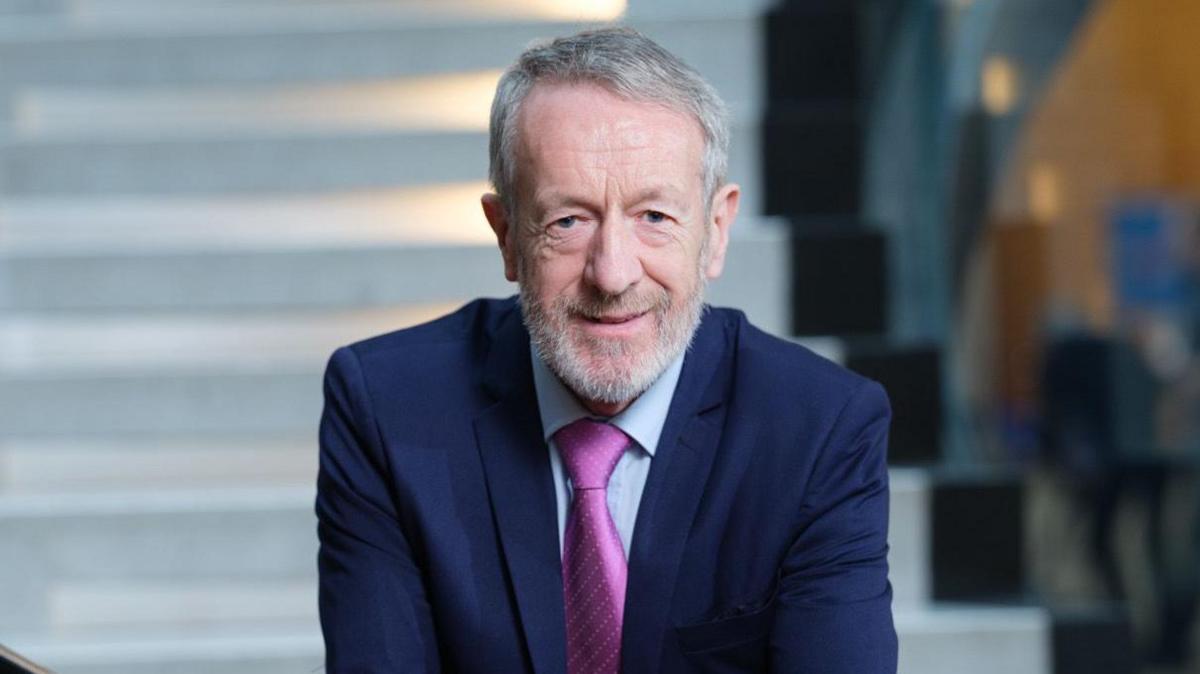 A grey short haired and bearded man wearing a navy suit and pink tie standing hands together on a bannister by some white stairs.