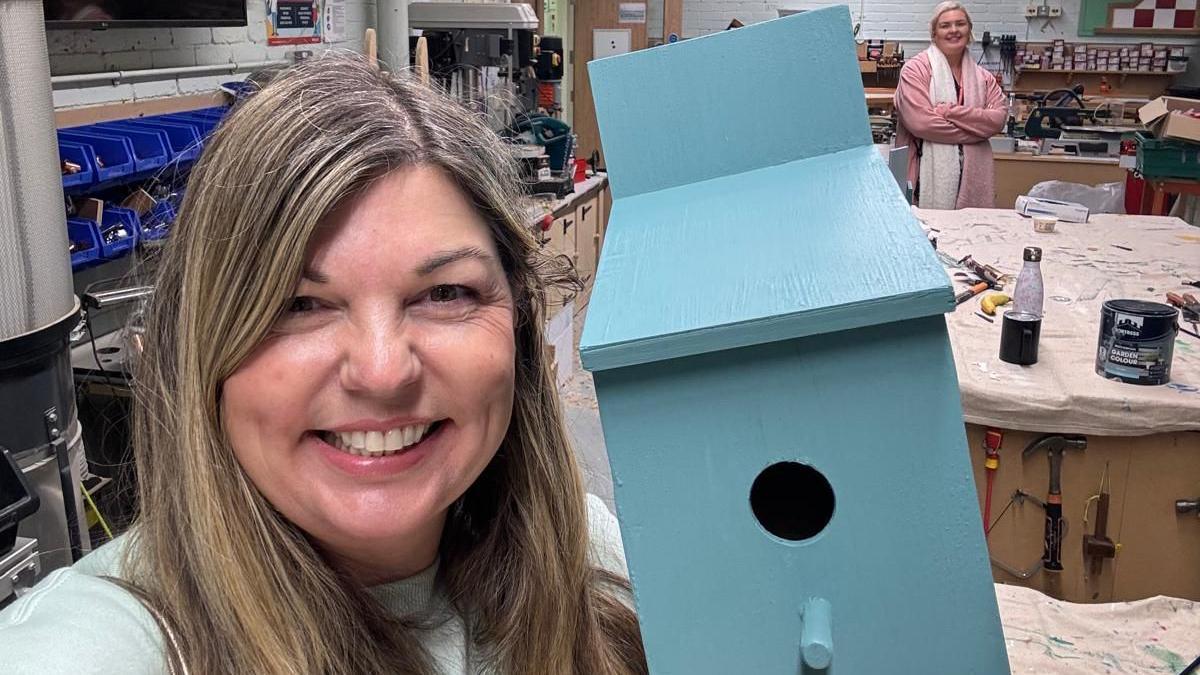 A smiling woman, with long blonde hair, takes a selfie of herself holding up a sky blue bird box, there are large work benches covered in dust sheets and paint cans in the background