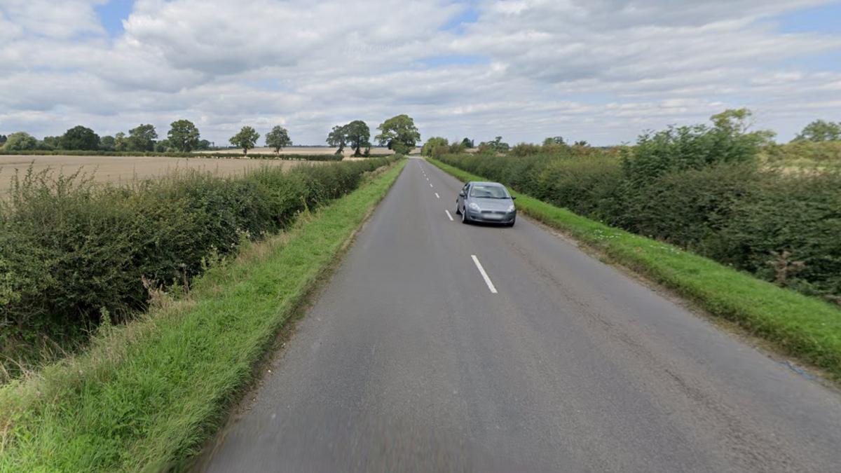 The B488 Leighton Road. A single grey car is travelling in the opposite direction and there are grass verges either side of the road