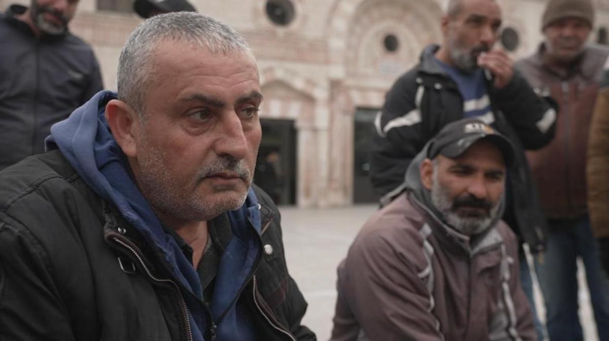 Two men sit next to each other in a square in Jordan with several men standing behind them