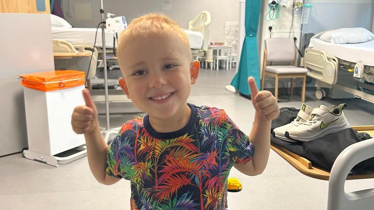 Harry smiles in a hospital in a brightly coloured palm print T-shirt. He has both thumbs up