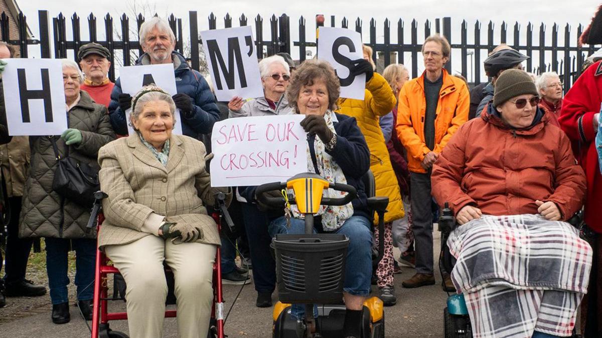 Protesters, including two women mobility scooters and a woman in a wheelchair, hold signs next to Wareham level crossing on 16 November 2024.